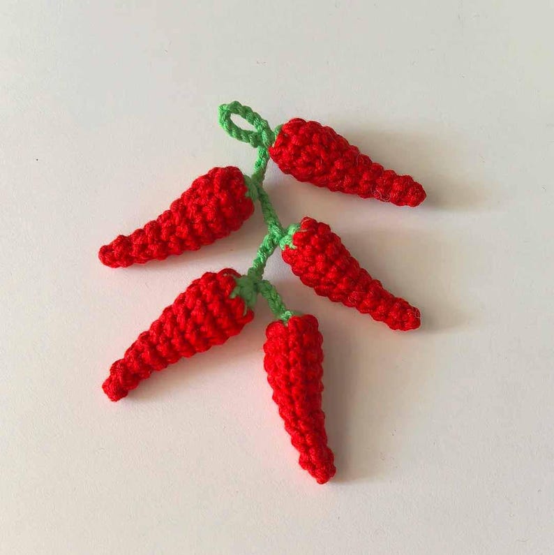 May include: A bunch of five crocheted red chili peppers with green stems. The peppers are arranged in a cluster, with the stems pointing upwards.