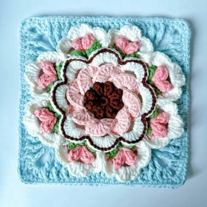 May include: A crocheted square with a large pink, white, and brown flower in the center. The flower has a scalloped edge and is surrounded by smaller pink flowers. The square is made with a light blue yarn.