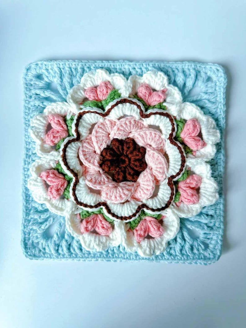 May include: A crocheted square with a large pink, white, and brown flower in the center. The flower has a scalloped edge and is surrounded by smaller pink flowers. The square is made with a light blue yarn.