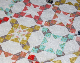 Puede incluir: Una colcha con fondo blanco y un patrón de estrellas en varios colores, incluyendo rojo, rosa, azul y amarillo. La colcha está hecha con una variedad de telas y tiene una textura acolchada.