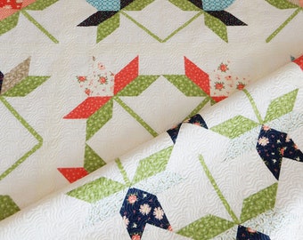 Puede incluir: Una colcha blanca con un patrón floral verde, azul y rojo. La colcha está hecha de cuadrados con un diseño en forma de estrella en el centro. La colcha está doblada por la mitad y el borde inferior es visible.