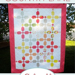 Może przedstawiać: Wzór patchworku o nazwie "Country Lane" autorstwa a Quilting Life Designs, z białym tłem i powtarzającym się wzorem kolorowych kształtów geometrycznych w odcieniach czerwieni, żółci, zieleni i pomarańczy. Patchwork jest zawieszony na sznurze do suszenia na świeżym powietrzu.