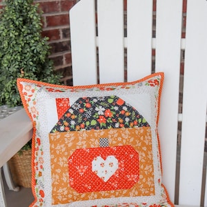 May include: A square quilted throw pillow featuring a central house-shaped design with a black floral roof and an orange pumpkin-shaped body. The pumpkin has a white heart in the center. The pillow is bordered by orange and white floral fabric.