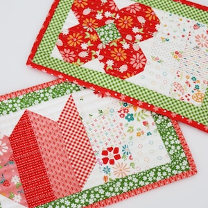 Pode incluir: Duas toalhas de mesa acolchoadas feitas à mão. Uma apresenta um desenho de flor vermelha com padrões florais e de morangos, e uma borda verde xadrez. A outra tem um padrão geométrico vermelho e branco com uma borda floral verde. Ambas têm uma guarnição vermelha.