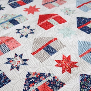 May include: A white quilt with a repeating pattern of red, white, and blue geometric shapes and star designs. The quilt features various fabrics with floral and patterned prints, creating a patriotic theme. The quilting pattern adds texture.