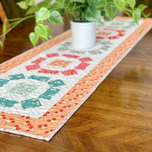 May include: A quilted table runner with a geometric pattern in shades of red, orange, white, and teal. The runner has a peach-coloured border with a floral print. The runner is on a wooden table with a potted plant in the background.