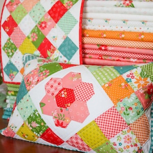 May include: A patchwork pillow with a large pink and red flower in the center. The pillow is made of various fabrics in shades of red, pink, green, and yellow. The pillow is sitting on a stack of fabric.