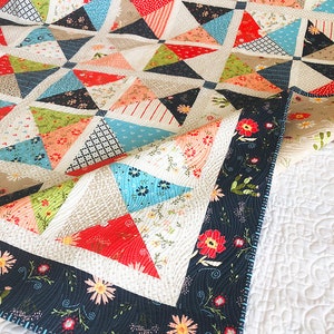 May include: A colorful quilt with a geometric design featuring triangles in red, blue, green, and floral patterns. The quilt has a dark blue border with floral and cactus motifs. The quilt is on a white, quilted surface.