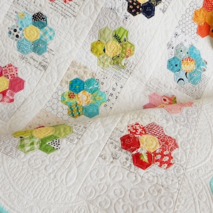 May include: A white quilt with a teal border. The quilt is made up of a patchwork of colorful hexagonal fabric flowers. The flowers are made of different fabrics and patterns. The quilt is quilted with a swirling pattern.