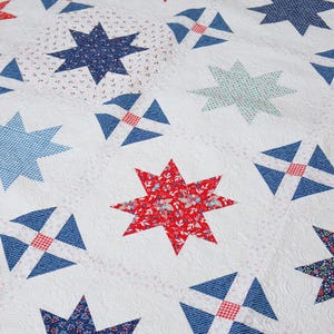 May include: A white quilt with a repeating pattern of red, blue, and light blue stars and geometric shapes. The stars are made of various patterned fabrics, including floral and gingham designs. The quilt has a quilted texture.