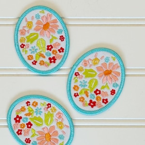 May include: Three oval-shaped embroidered patches featuring a white background with a colorful floral pattern of pink, red, yellow, and light blue flowers and green leaves, all bordered by a bright turquoise stitched edge.