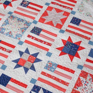 May include: A white quilt featuring a repeating pattern of American flags and stars in red, white, and blue. The quilt has a quilted texture with intricate stitching, creating a patriotic theme. The flags and stars are made of various patterned fabrics.