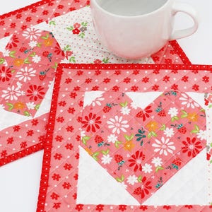May include: A white ceramic mug rests on a quilted heart-shaped coaster. The coaster features a red floral pattern with a white heart center. A second coaster with a similar design is also visible.