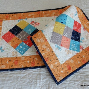 May include: Two quilted table runners with a geometric patchwork design in shades of orange, blue, red, and gray. The runners have a white background with a floral pattern and a navy blue border.