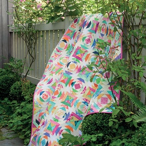 Op de afbeelding: Een kleurrijk patchwork-deken met een geometrisch ontwerp, gemaakt van fel roze, blauw, groen en geel stof. Het deken is over een wit schuttingshek in een tuin gelegd.