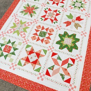 May include: A handmade quilt featuring a patchwork design with red, green, and white star patterns. The quilt has a red floral border and is laid out on a neutral-colored surface. The quilt is a festive, decorative item.