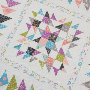 May include: A detailed close-up of a handmade quilt with a starburst design. The quilt features a white background with colorful triangles in shades of purple, green, blue, and peach. Floral patterns and decorative stitching add to the quilt's charm.