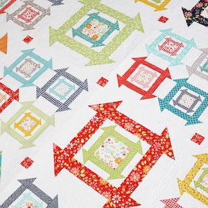 May include: A white quilt with a repeating pattern of colorful squares with arrows pointing in different directions. The squares are made of various fabrics with floral and geometric patterns.