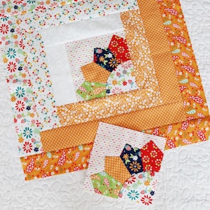 May include: A colorful patchwork quilt with a log cabin design. The quilt features a central block with a fan-like pattern in red, navy, orange, and green. The quilt is surrounded by orange and floral patterned fabric. A smaller, matching block is also present.