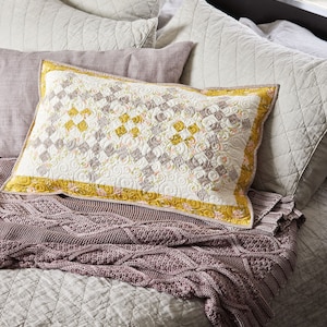 May include: A bed with a white quilted bedspread and a purple knitted throw blanket. There are several pillows on the bed, including a patterned pillow with a yellow border.