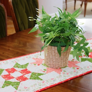 Pode incluir: Um corredor de mesa de patchwork branco, vermelho e verde com uma borda de padrão floral. Uma planta em vaso com folhas verdes e flores brancas está sobre o corredor de mesa.