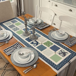 May include: A dining table set for a meal, featuring a quilted table runner with blue, green, and white square patterns. The table is set with white plates, silverware, and gray placemats. Two deer-shaped candle holders and candles are also on the table.