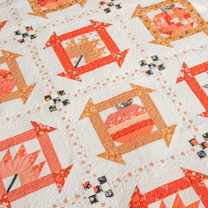 Pode incluir: Um edredom feito à mão com um padrão repetido de folhas e abóboras laranja e enferrujadas, emolduradas em quadrados com detalhes de setas. O edredom tem um fundo branco com detalhes florais e geométricos.