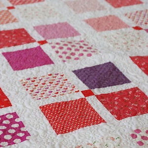 Può includere: Una trapunta con un motivo ripetuto di quadrati colorati nelle tonalità del rosso, rosa e viola. I quadrati sono su uno sfondo bianco con trapuntatura floreale.