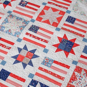 May include: A patriotic quilt featuring a repeating pattern of American flags and stars in red, white, and blue. The quilt has a white background with intricate quilted details. The flags and stars are made from various patterned fabrics.
