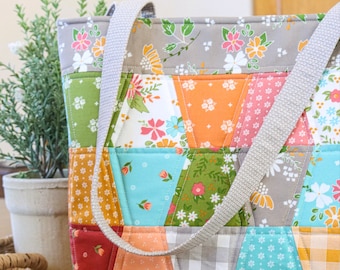 Könnte beinhalten: Eine Patchwork-Tragetasche mit verschiedenen floralen und geometrischen Mustern in Grün, Orange, Rosa und Grau. Die Tasche hat einen grauen Riemen und steht auf einem Weidenkorb. Es ist ein handgefertigter Artikel.