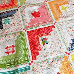Peut inclure: Une courtepointe patchwork colorée avec une variété de motifs et de formes. La courtepointe présente des carrés avec des motifs de cabanes en rondins, des étoiles et des motifs floraux dans des tons de rouge, vert, orange et bleu. La courtepointe est en tissu.