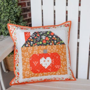 Peut inclure: Un coussin décoratif matelassé carré présentant un motif en patchwork central représentant une citrouille avec un cœur blanc au milieu. La citrouille est orange et rouge avec des motifs floraux, surmontée d'un toit floral sombre et d'un passepoil orange.