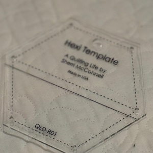 May include: Clear acrylic Hexi Template with the text "Hexi Template" and "A Quilting Life by Sherri McConnell Made in USA". The template has a hexagonal shape with dotted lines and the text "QLD-R01".