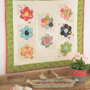 Peut inclure: Une courtepointe avec un fond blanc et une bordure verte. La courtepointe est composée de plusieurs formes de fleurs hexagonales de différentes couleurs, notamment le rouge, le bleu, le jaune et le vert. La courtepointe est accrochée à un mur blanc au-dessus d'une table en bois avec un plat blanc et un pot gris avec des fleurs roses.