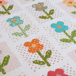 May include: A white quilt with a repeating pattern of colorful flowers with green stems and leaves. The flowers are made from different patterned fabrics and are arranged in a grid pattern.