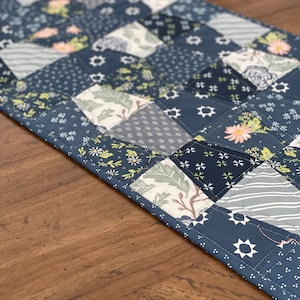 May include: A blue and white patchwork table runner featuring a variety of floral and geometric patterns. The runner is made from fabric squares sewn together in a diagonal pattern.