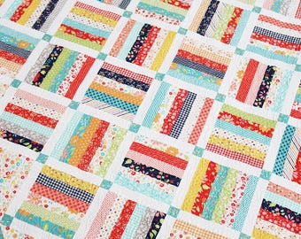 Puede incluir: Un edredón de patchwork colorido con fondo blanco. El edredón presenta bloques rectangulares con tiras de varias telas estampadas en rojo, amarillo, azul y verde. El borde tiene un estampado floral.