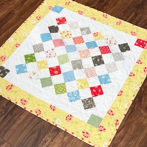 Może przedstawiać: Biała kołdra z żółtą obwódką i wzorem kwiatowym. Kołdra składa się z małych kwadratów w różnych kolorach i wzorach, w tym czerwonego, niebieskiego, zielonego i szarego.