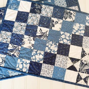 May include: Two blue and white patchwork placemats with a quilted design. The placemats are made of various blue and white floral fabrics.