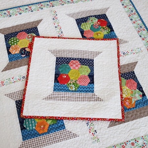 May include: A square quilted wall hanging featuring a central spool of thread motif made from colorful hexagonal fabric patches. The spool is surrounded by a white background and a red border. A larger matching quilt lies underneath the smaller piece.
