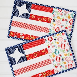May include: Two patriotic placemats with a patchwork design. One side features a blue field with a white star, red and white stripes, and a red and white gingham pattern. The other side has a floral pattern with red, pink, and yellow flowers.
