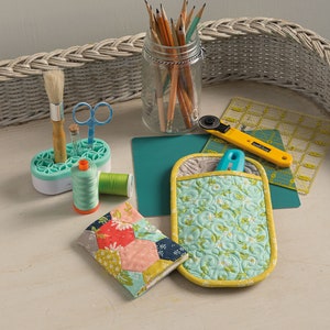 May include: A white wicker tray with a variety of craft supplies, including a jar of pencils, a pair of scissors, a paintbrush, a spool of thread, a small sewing project, a rotary cutter, a ruler, and a quilted hot pad.