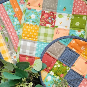 May include: Two colorful patchwork pot holders with a blue and white checkered pattern, a floral pattern, and a green and white plaid pattern. The pot holders are on a wooden surface with green leaves.