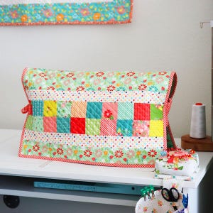 May include: A quilted sewing machine cover with a patchwork design. The cover features a mix of colorful squares and floral patterns in red, green, yellow, and blue. The edges are trimmed with a red and white gingham fabric. A matching wall hanging is visible in the background.