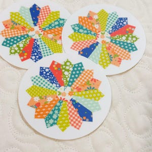 May include: Three white, circular coasters with a colorful, patchwork flower design. Each coaster features a vibrant arrangement of fabric patterns in shades of blue, green, orange, and red. The coasters are set against a white, quilted background.