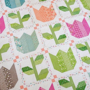 Könnte beinhalten: Ein Quilt mit einem sich wiederholenden Muster aus Tulpenblöcken in verschiedenen Farben wie Pink, Orange, Blaugrün und Braun, jeweils mit grünen Stielen und Blättern. Die Blöcke sind durch weißen Stoff mit kleinen Blumenakzenten getrennt.