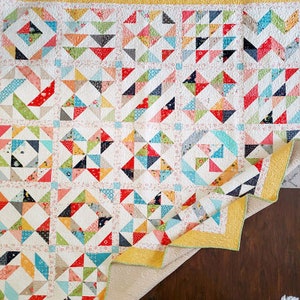 May include: A colorful patchwork quilt with a white background and a yellow border. The quilt is made up of various geometric shapes, including squares, triangles, and diamonds. The quilt is folded in half and is lying on a wooden surface.