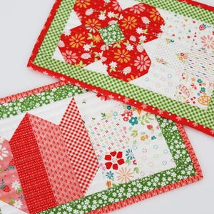 May include: Two handmade quilted placemats. One features a red flower design with floral and strawberry patterns, and a green gingham border. The other has a red and white geometric pattern with a green floral border. Both have a red trim.