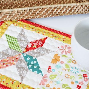 May include: A quilted star-shaped placemat with a floral and geometric pattern. The placemat features a starburst design in shades of green, red, gray, and pink, with a yellow floral border and a red dotted edge. A white ceramic mug is partially visible.