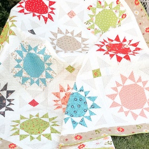 May include: A white quilt with a patchwork design featuring various colored starburst patterns. The quilt is made with different fabrics, including floral prints, geometric patterns, and solid colors. The quilt is folded and draped over a white wicker chair.